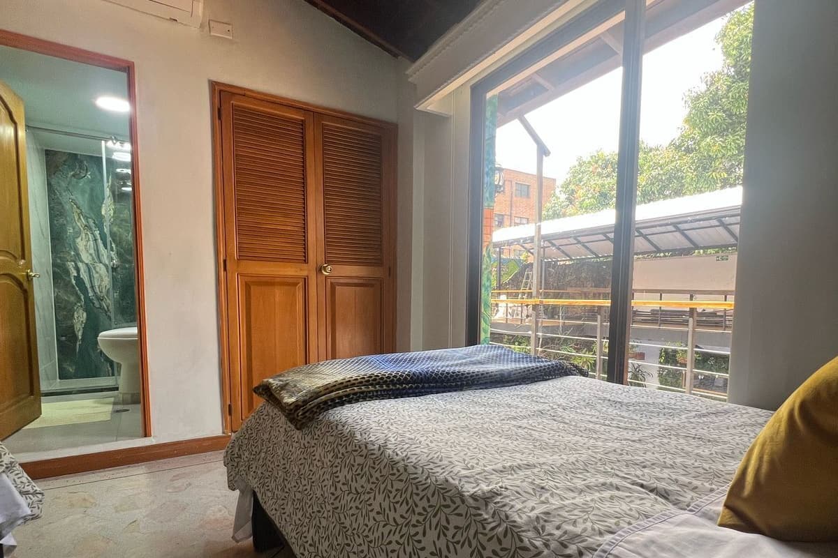 A bedroom with a bed, a wooden wardrobe, a bathroom with a glass door, and a large window with a view of the outside.