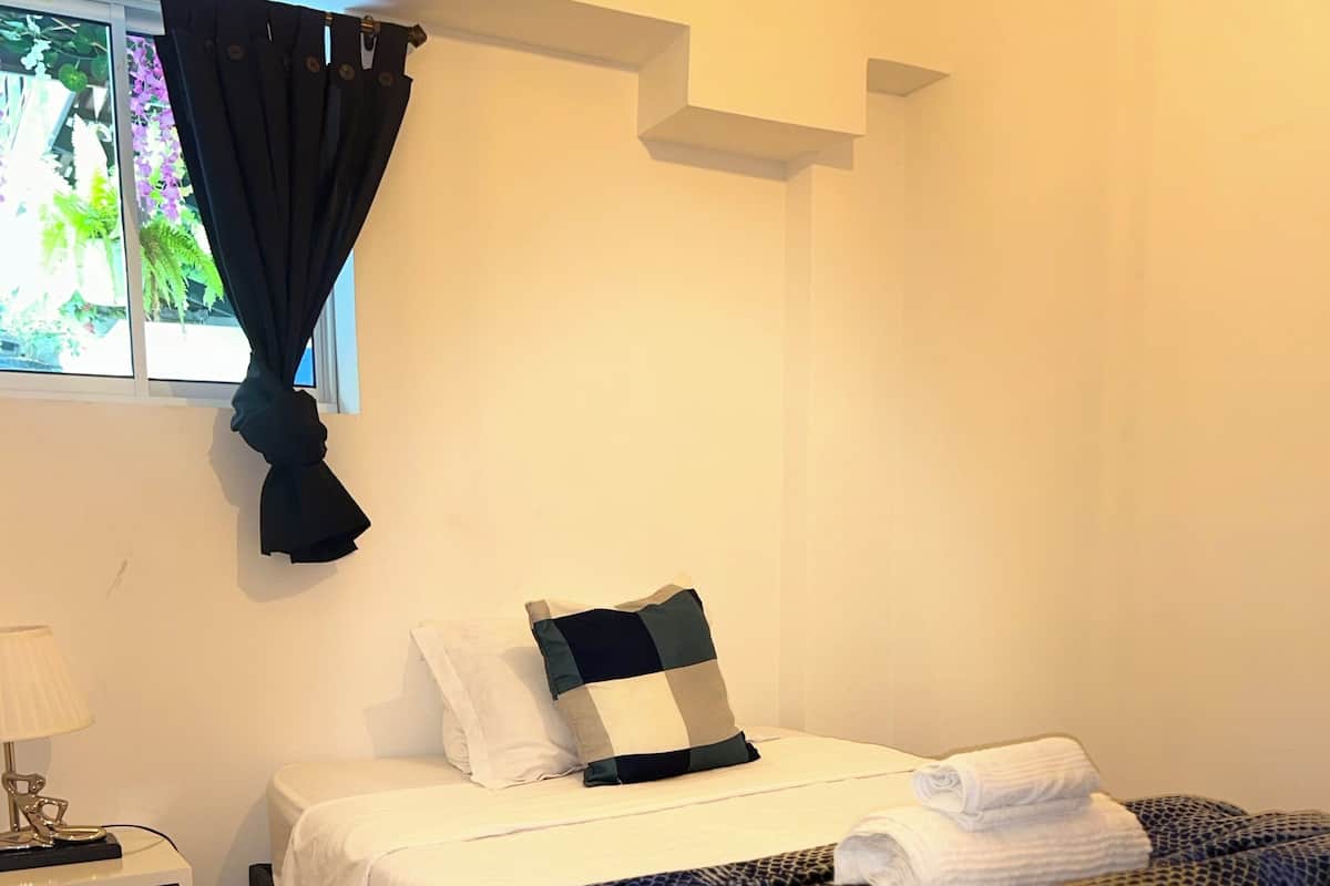 A bedroom with a bed, a nightstand, a window with curtains, and a view of greenery outside.