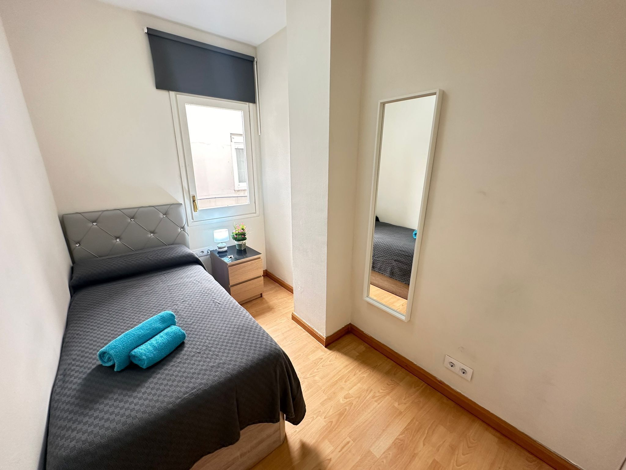 Un dormitorio con una cama, una mesita de noche, una ventana con cortina y un espejo en la pared.