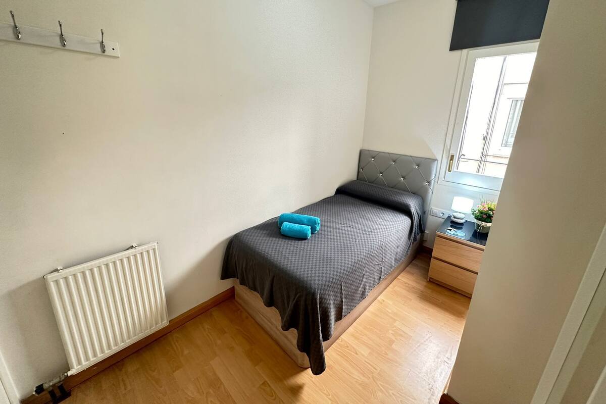 A small bedroom with a bed, a radiator, a window with a view, and a bedside table with a plant.
