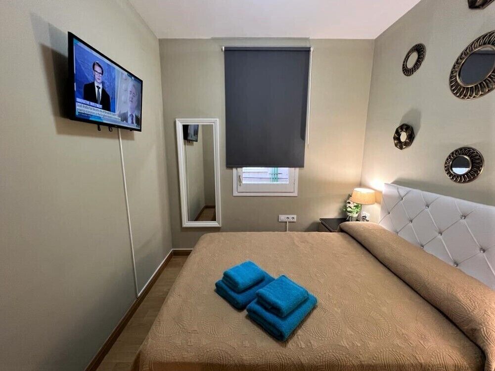 Habitación de hotel con una cama, televisor montado en la pared, ventana con persiana y espejos decorativos.