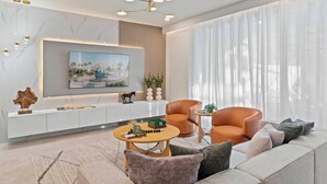 Een moderne woonkamer met een grote flatscreen televisie bevestigd aan een witte console, een ronde salontafel, een leren fauteuil en een bank met meerdere kussens.