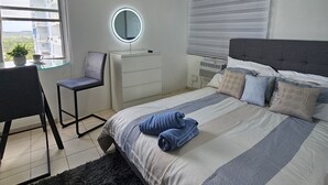 Quarto com cama, escrivaninha, cadeira, espelho redondo e janela com persianas.
