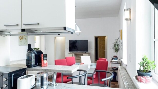 A modern kitchen with a built-in stove, a coffee maker, and a toaster. There is a dining area with red chairs and a table. A TV is mounted on the wall, and a plant is on the windowsill. - Beautiful living near Schönbrunn (Wien)