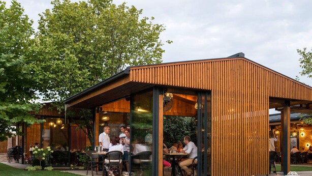 Un espace extérieur aménagé avec des tables et des chaises, entouré d’une structure en bois aux murs de verre, sous un ciel dégagé.
