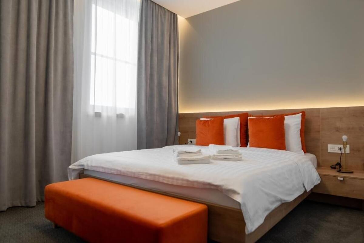 Ein Hotelzimmer mit Bett, einer orangefarbenen Bank, einem Nachttisch und einem Fenster mit Vorhängen.