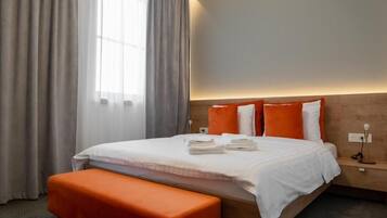 A hotel room with a bed, an orange bench, a nightstand, and a window with curtains.