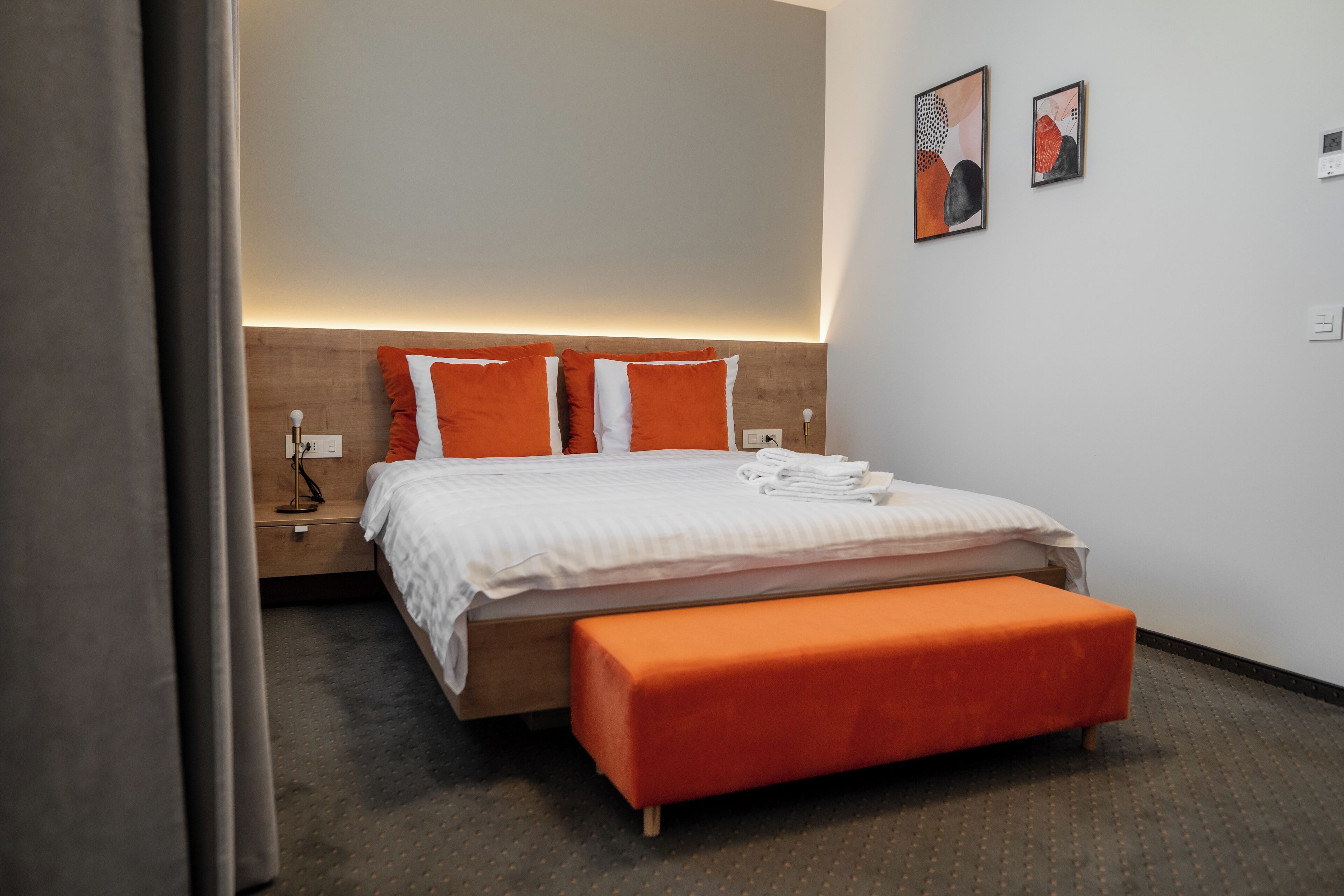 Une chambre d’hôtel moderne avec une tête de lit en bois, un grand lit avec une literie orange, un banc assorti et deux œuvres d’art accrochées au mur.