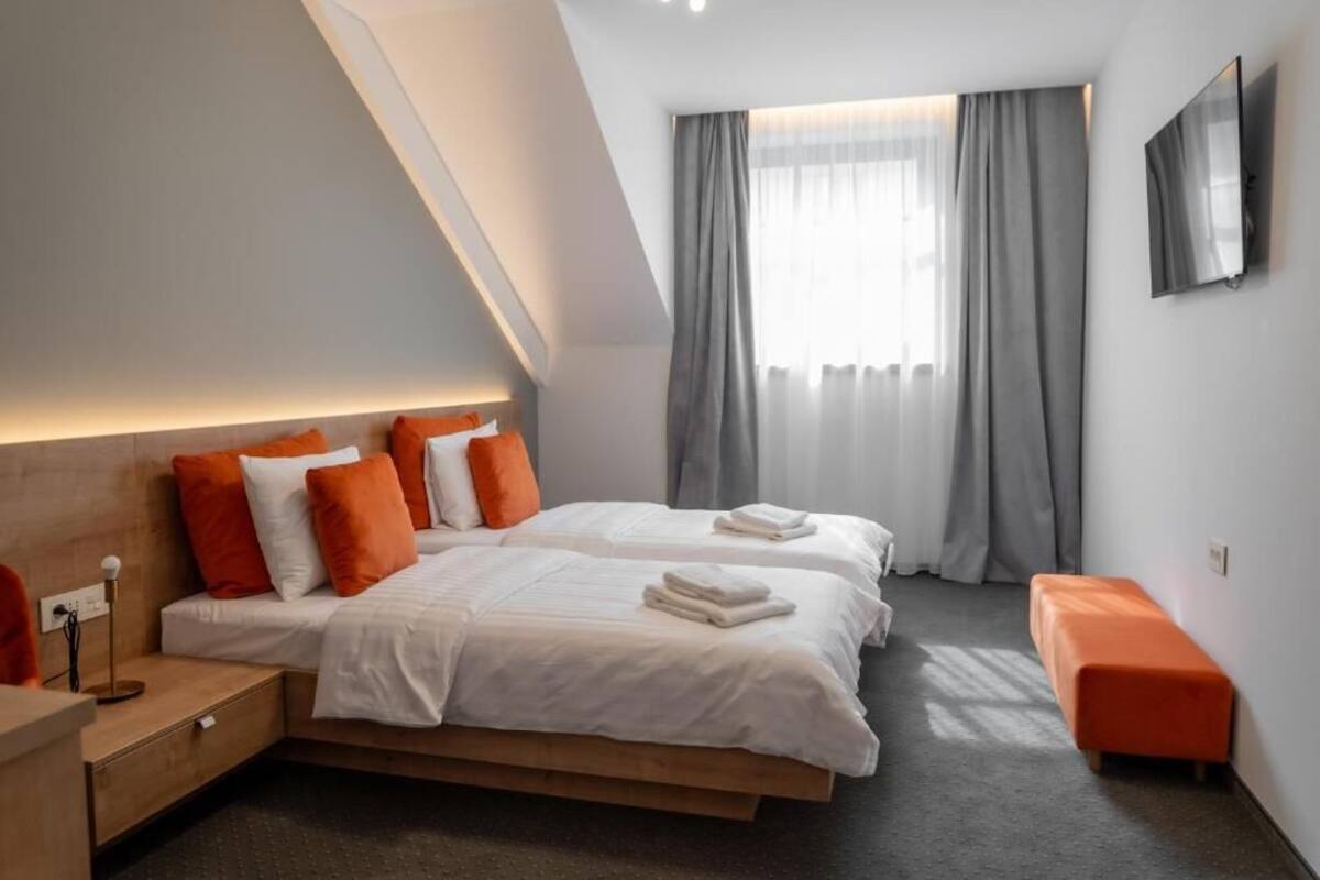 Ein modernes Hotelzimmer mit zwei Betten, einem Holzkopfende, einer Bank, einem Fernseher und einem großen Fenster mit Vorhängen.