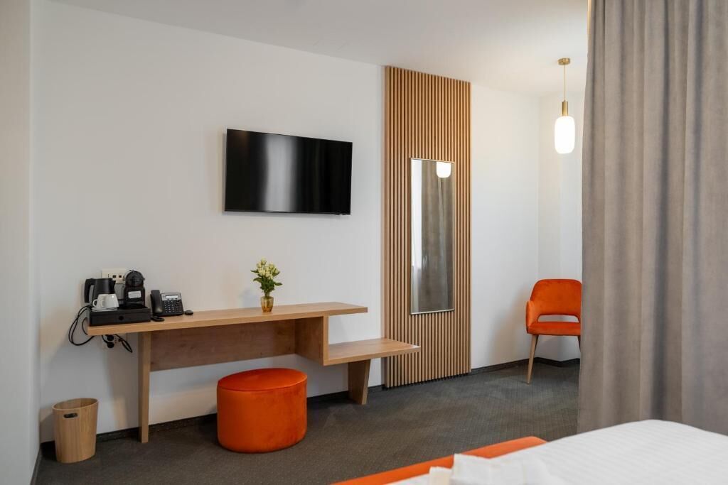 A hotel room with a bed, a desk with a flat-screen TV, a small orange stool, a wooden panel wall, and a single orange chair.
