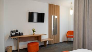 A hotel room with a bed, a desk with a flat-screen TV, a small orange stool, a wooden panel wall, and a single orange chair.