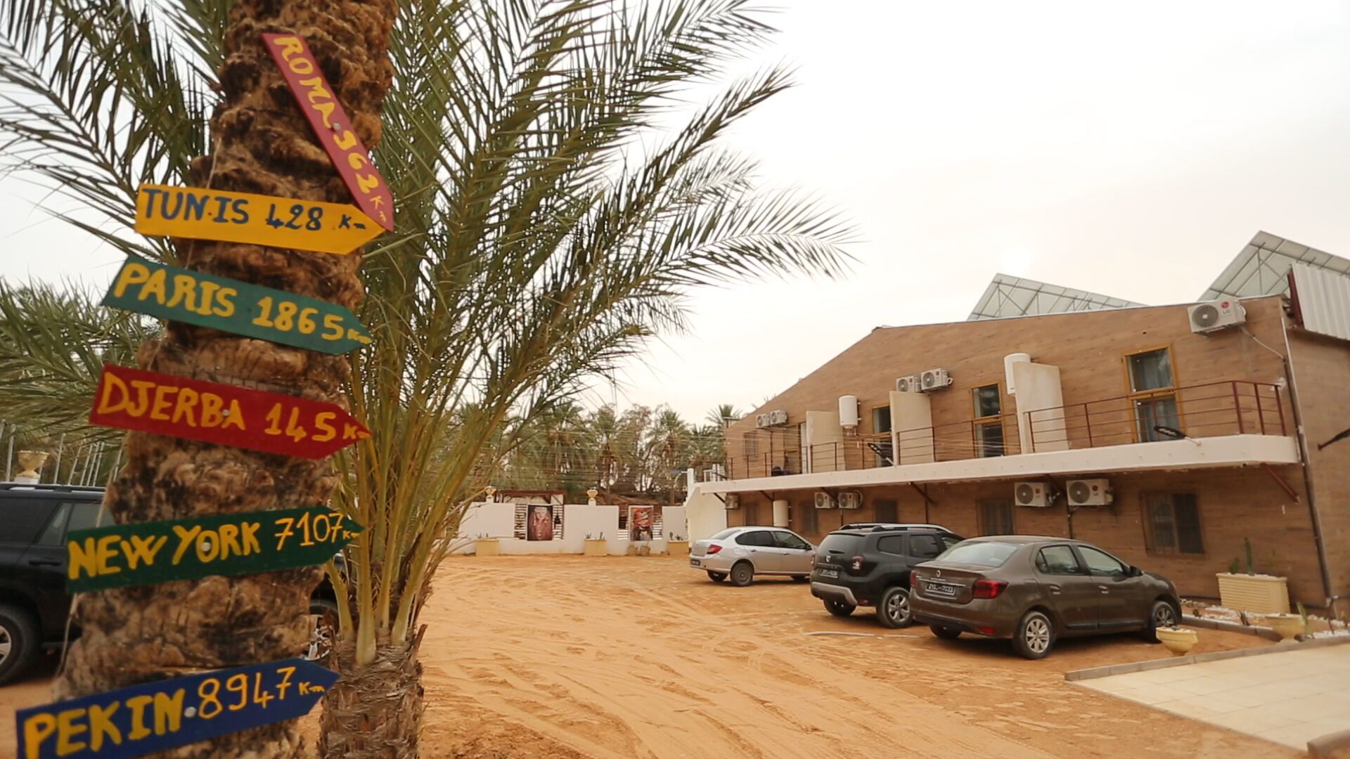 A building with multiple air conditioning units on the roof, parked cars, and a palm tree with directional signs.