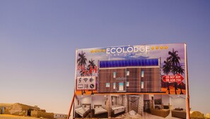 Un letrero de hotel que anuncia servicio las 24 horas, números de contacto y un edificio moderno con paneles solares.