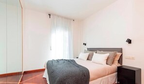 A bedroom with a bed, a nightstand, a window with curtains, and a wall-mounted light.