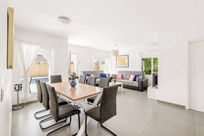 A modern living room with a dining area, a grey sofa set, a TV, and a pool visible through the glass doors. - Villa Esmerelda Sun - Four Bedroom Villa, Sleeps 8 (Bel-Air)