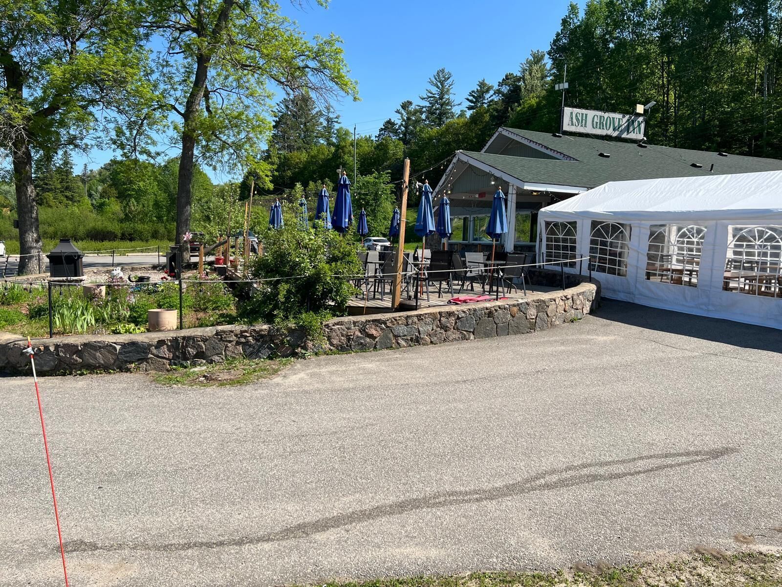 A covered outdoor seating area with tables and blue umbrellas, a stone retaining wall, and a building labeled "ASH GROVE".
