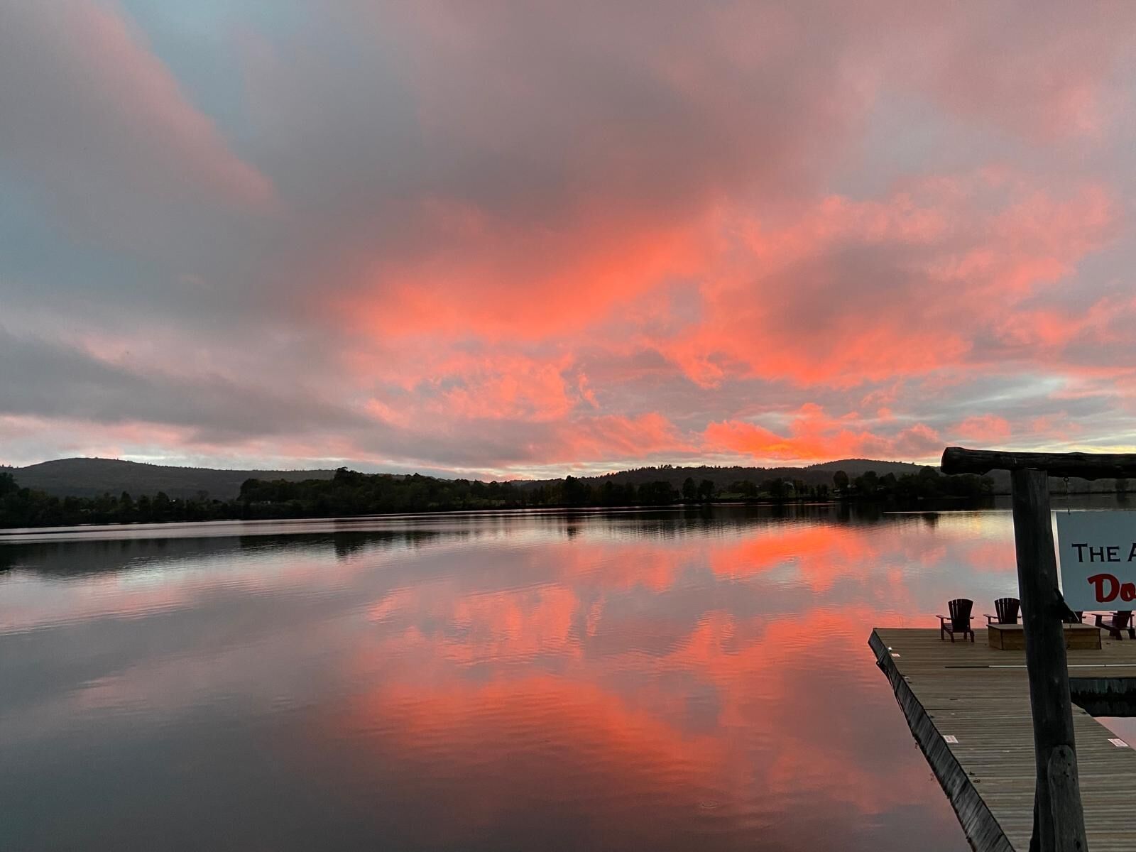 A calm lake with a wooden dock and chairs, surrounded by hills.