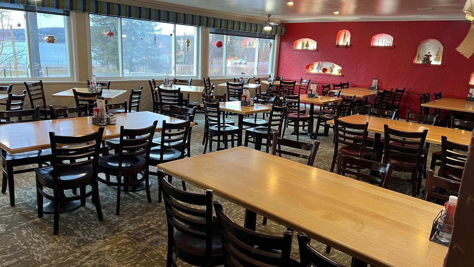 A restaurant with multiple wooden tables and chairs, large windows with decorations, and a red wall with small windows.