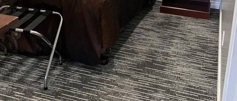 Habitación de hotel con cama, silla de ruedas, mesita de noche, lámpara y un cuadro de un lago con follaje otoñal.