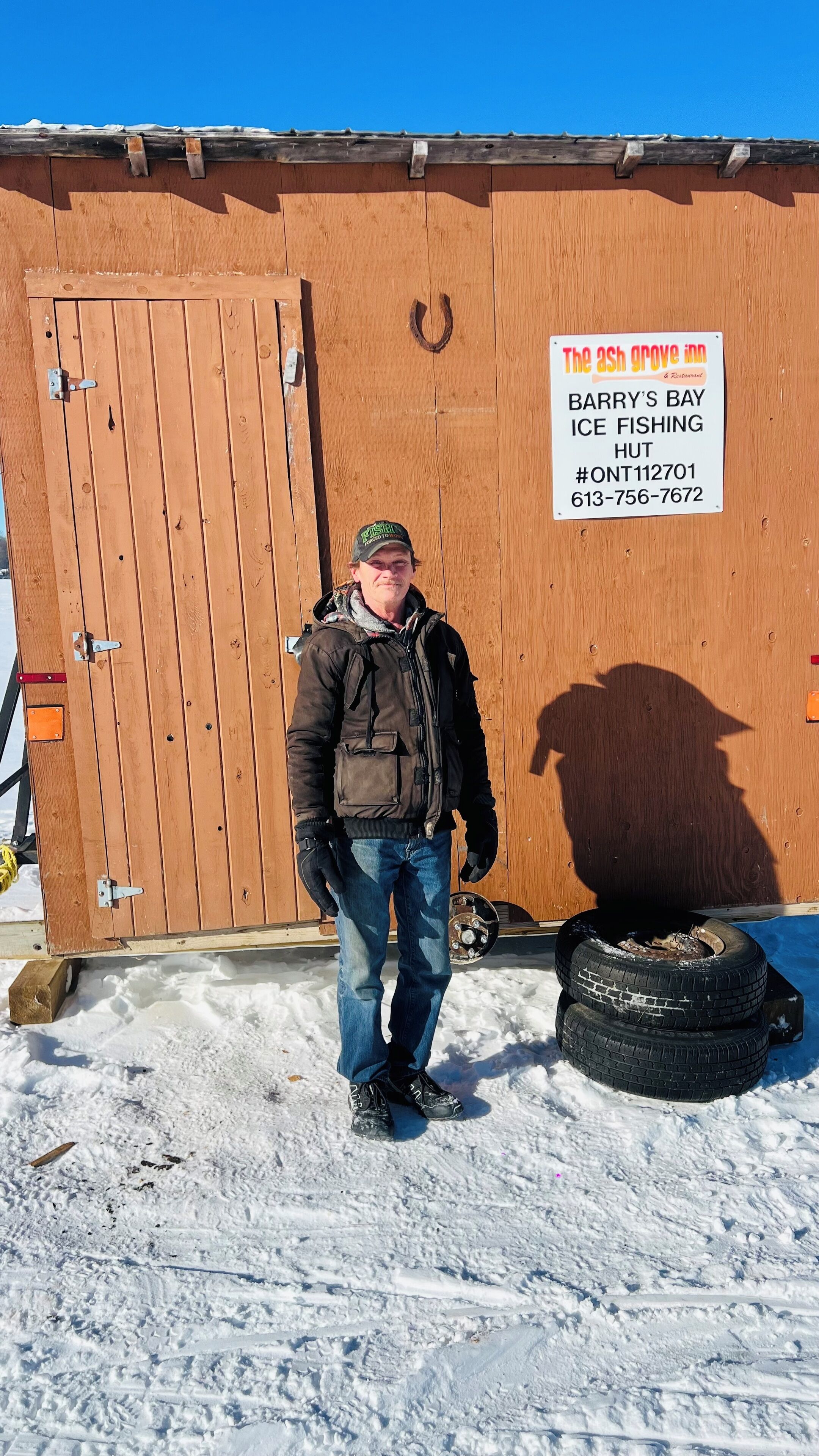 Un homme se tient devant un bâtiment en bois sur lequel est apposée une enseigne indiquant « The Ash Grove Inn » et mentionnant « Barry's Bay Ice Fishing Hut ».