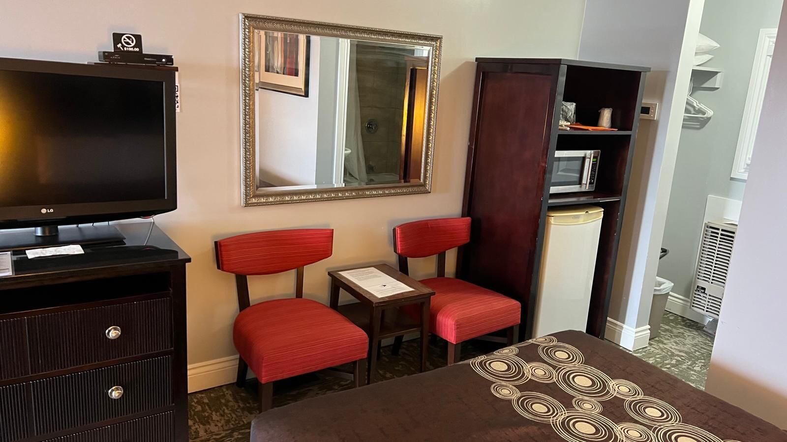 A hotel room with a flat-screen TV, a mirror, two red upholstered chairs, a small table, and a bed with a patterned bedspread.