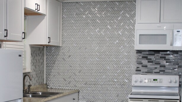 Una cocina con gabinetes blancos, microondas, horno, pileta y salpicadero con motivo de espiga.