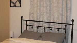 Una cama con estructura metálica, una colcha color beige y dos almohadas. Junto a la cama, una mesita de noche con una lámpara.