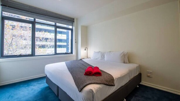Una habitación de hotel con una cama grande, una ventana con persianas, una mesita de noche y una alfombra azul.