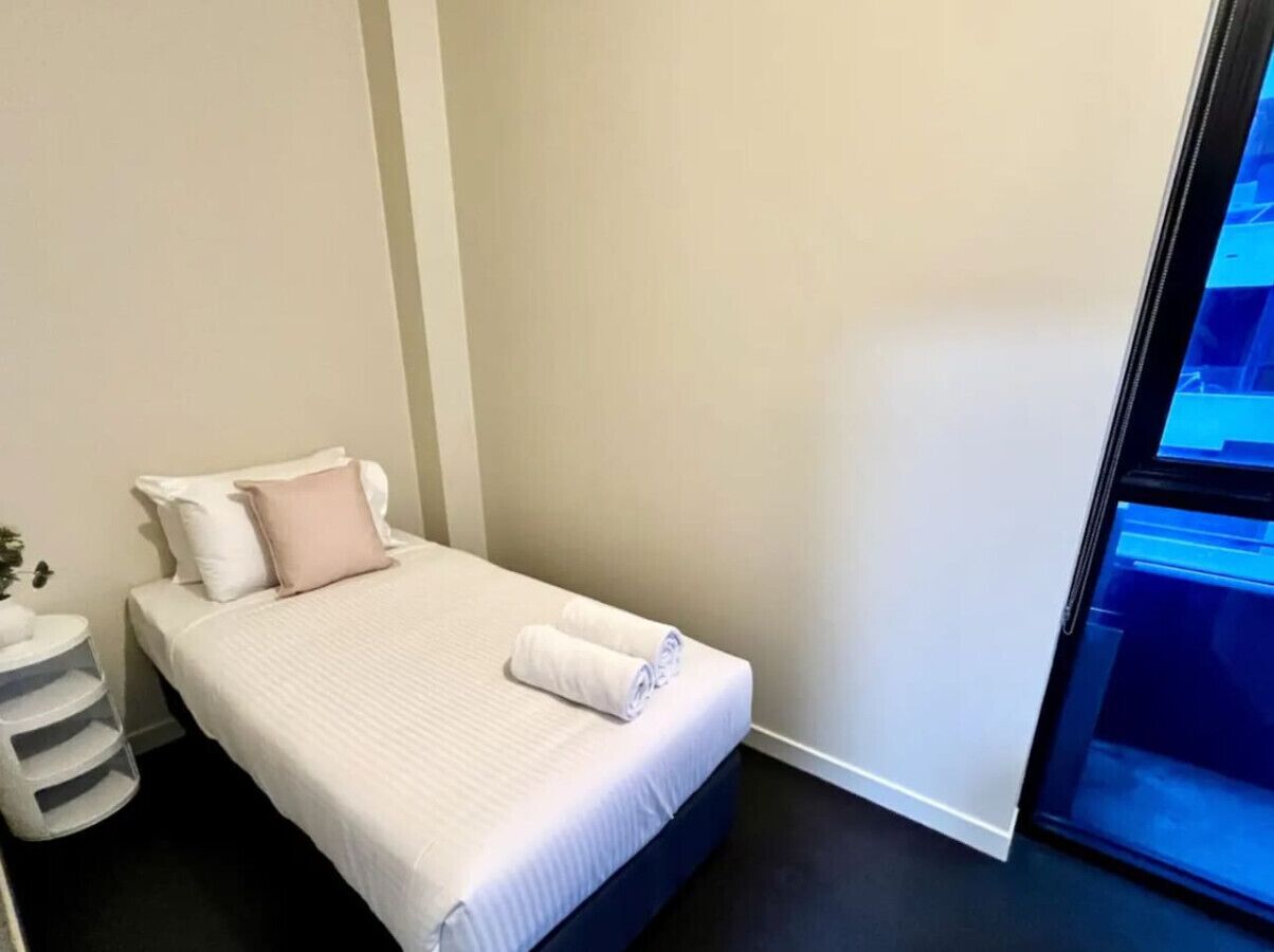 Een enkel bed met witte beddengoed en een roze kussen, een klein nachtkastje met een plant, en een raam met uitzicht op een blauwe lucht.
