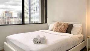 A modern hotel room with a large bed, a view of the cityscape through a large window, and a neatly made bed with rolled towels. - Lonsdale Heights (Higher Floor) 1BR with City View (Melbourne)