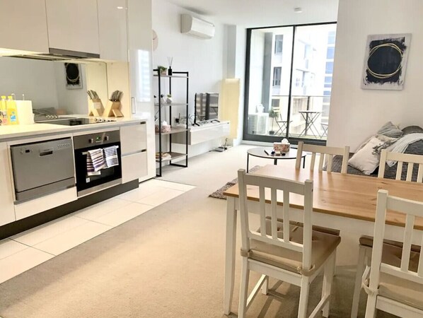 A modern kitchen with white cabinetry, a dining area with a wooden table and white chairs, and a view of the city through large windows.