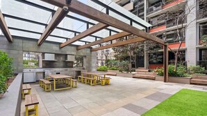Ein Terrassenbereich im Freien mit mehreren Holztischen und Bänken, einer Rasenfläche und Blick auf ein modernes Gebäude mit Balkonen.