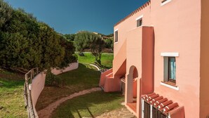 A building with a pink facade, a stone pathway, and a view of the surrounding greenery and hills. - Apartments With Pool Country Village (Stintino)