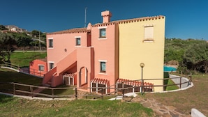 A two-story building with a red and yellow facade, a staircase, and a pool area. - Apartments With Pool Country Village (Stintino)