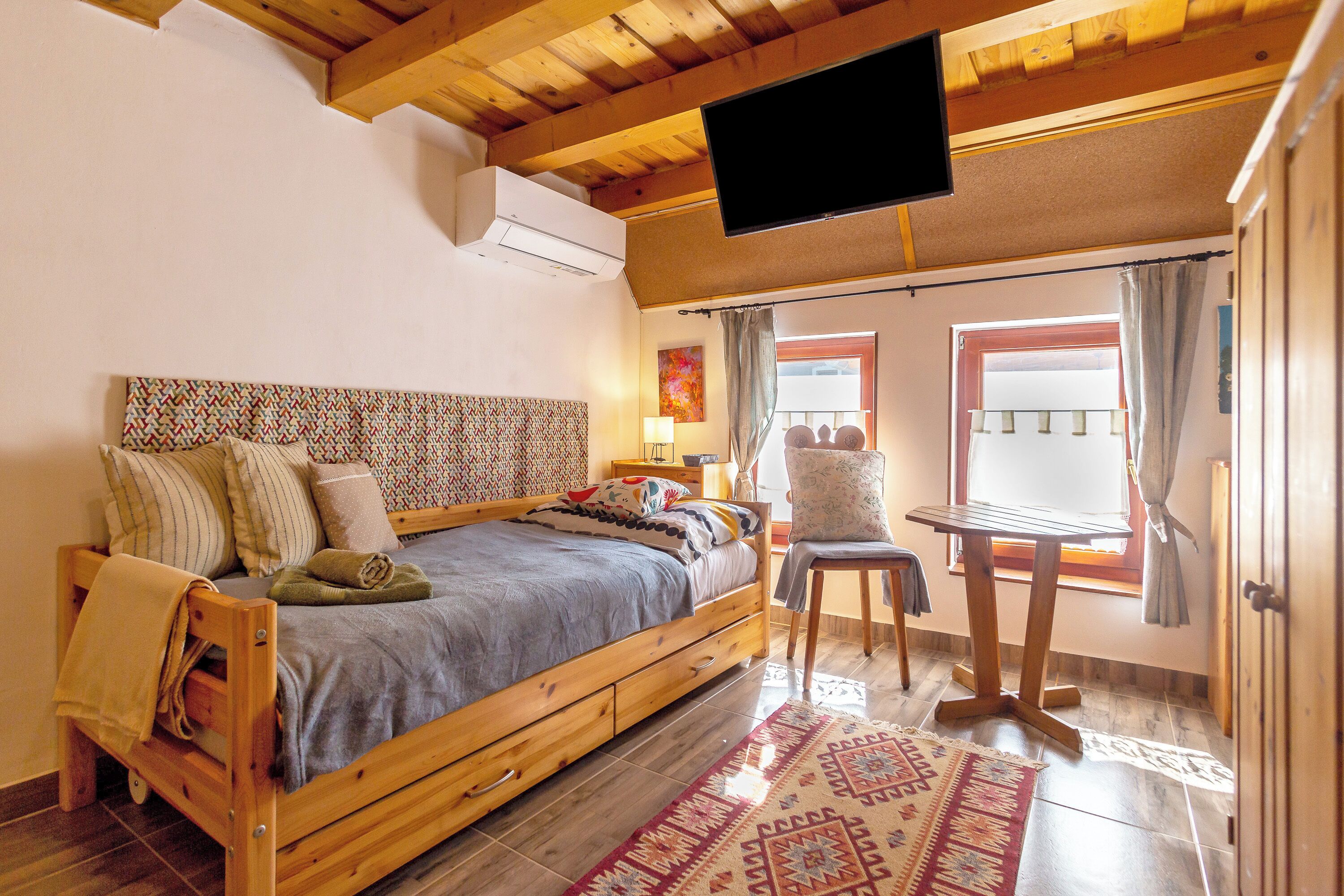 Un llit de fusta amb un capçal estampat, un televisor fixat al sostre, una taula petita i una finestra amb cortines.