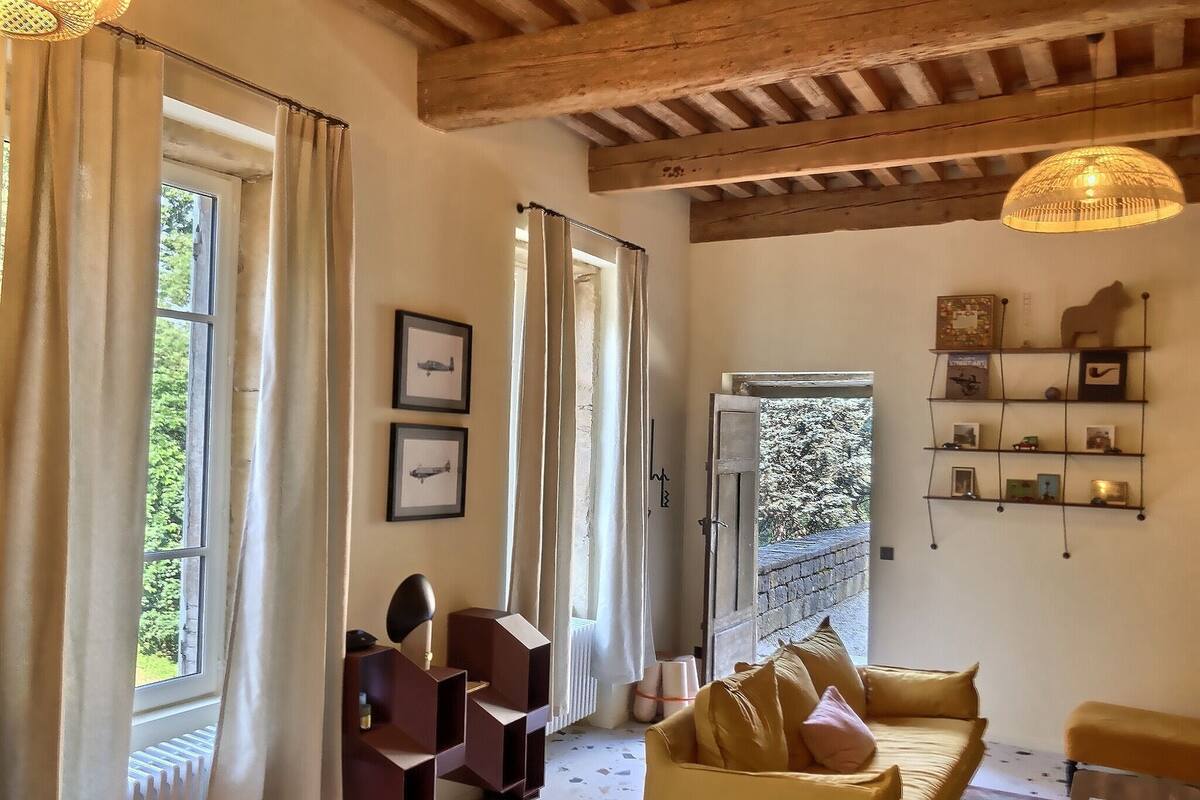 A cozy living room with a yellow sofa, wooden beams on the ceiling, and a view of greenery outside.