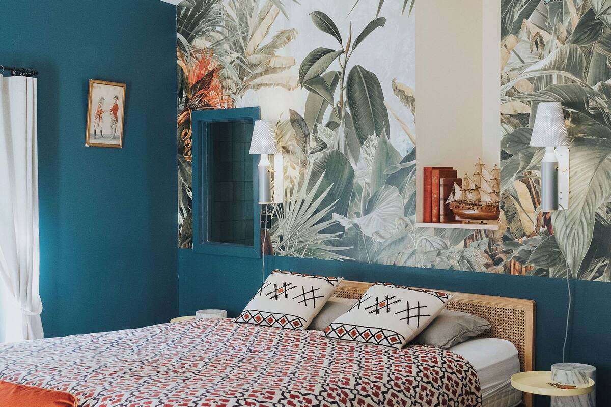 A bedroom with a teal wall, a patterned bedspread, a wooden headboard, a small bedside table, and a mirror.