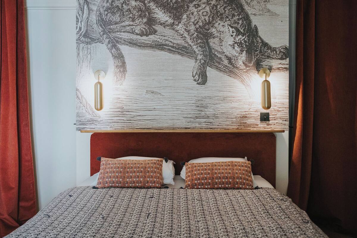 A bed with a patterned bedspread, two patterned pillows, a headboard with a wooden panel, and a wall-mounted light fixture.