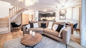 A modern living room with a wooden staircase, a sectional sofa, a round coffee table, and a kitchen in the background. - Gentiana - Cosy apartment in Les Gets (Les Gets)