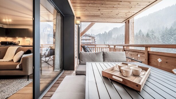 A wooden deck with a table and chairs, a sofa, and a sliding glass door leading to a kitchen. - Gentiana - Cosy apartment in Les Gets (Les Gets)