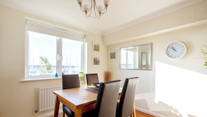 Un comedor con una mesa de madera y cuatro sillas, un ventanal con vistas a barcos, un reloj de pared y cuadros enmarcados.