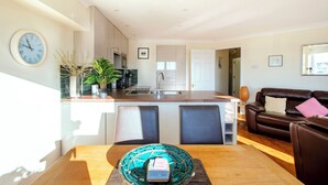 Una cocina moderna con una isla central, un comedor con mesa y sillas, y una sala de estar con sofá y cojines.