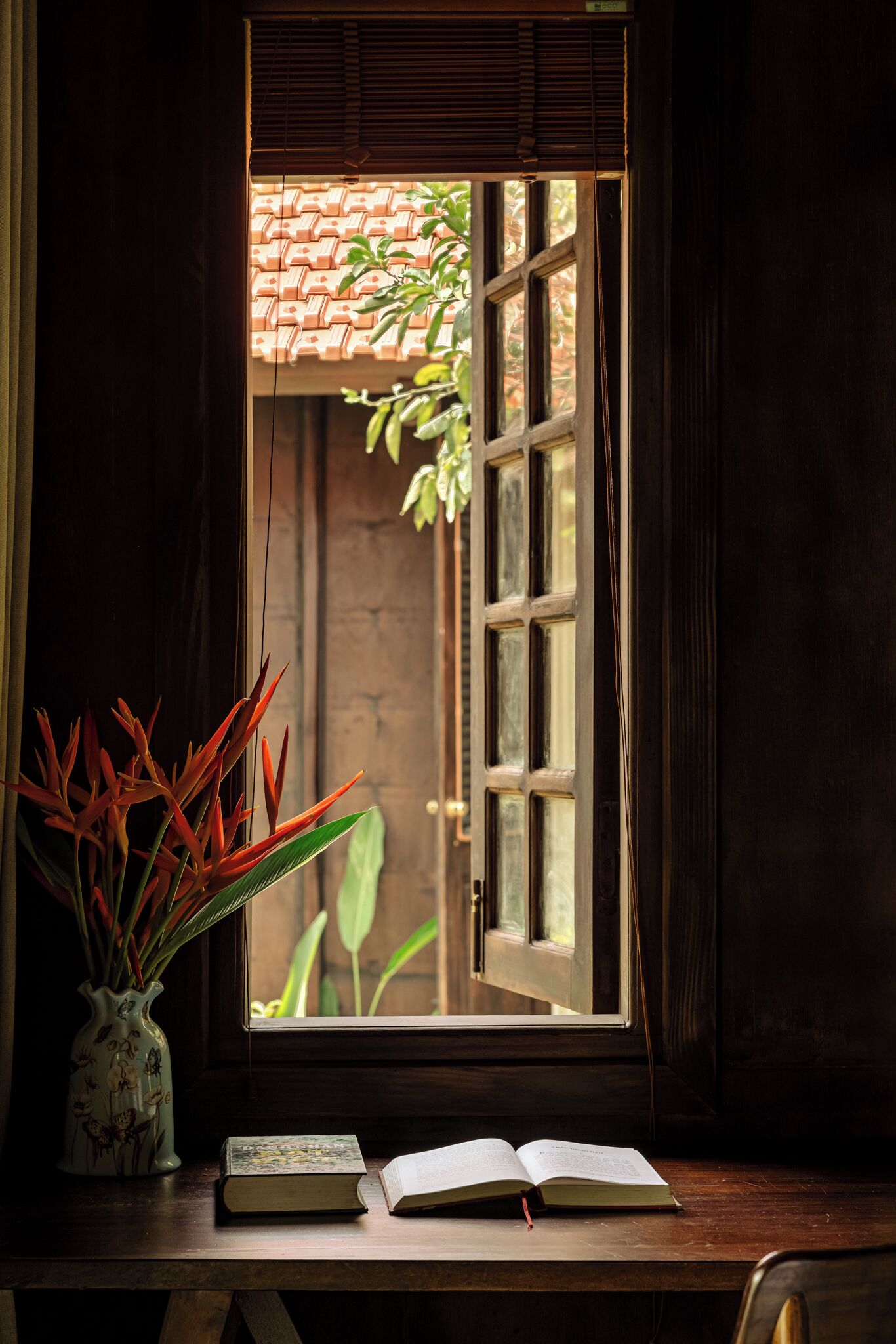 A window with a view of a tiled roof and plants, a vase with flowers, and an open book on a wooden table.