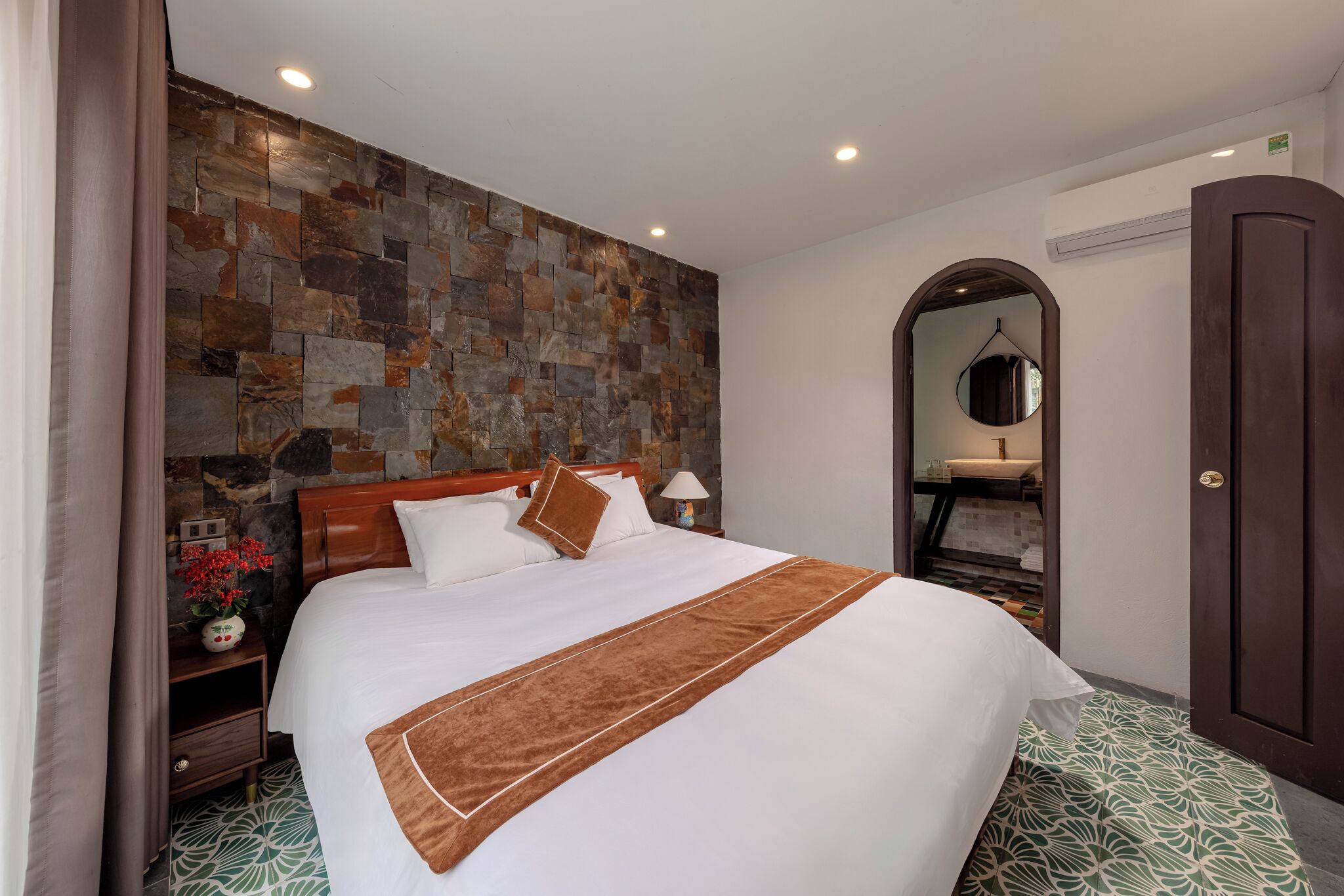 A neatly made bed with a brown accent, a stone wall, a mirror, and a door.