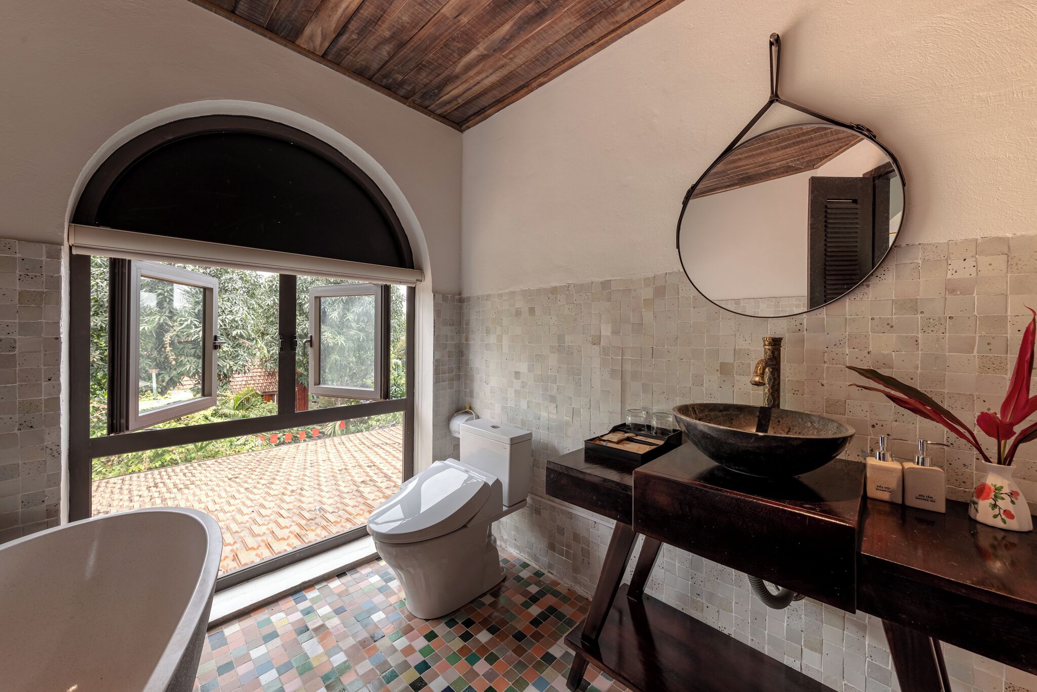 Une salle de bain avec une grande fenêtre, un miroir rond, un lavabo avec un receveur et des toilettes.