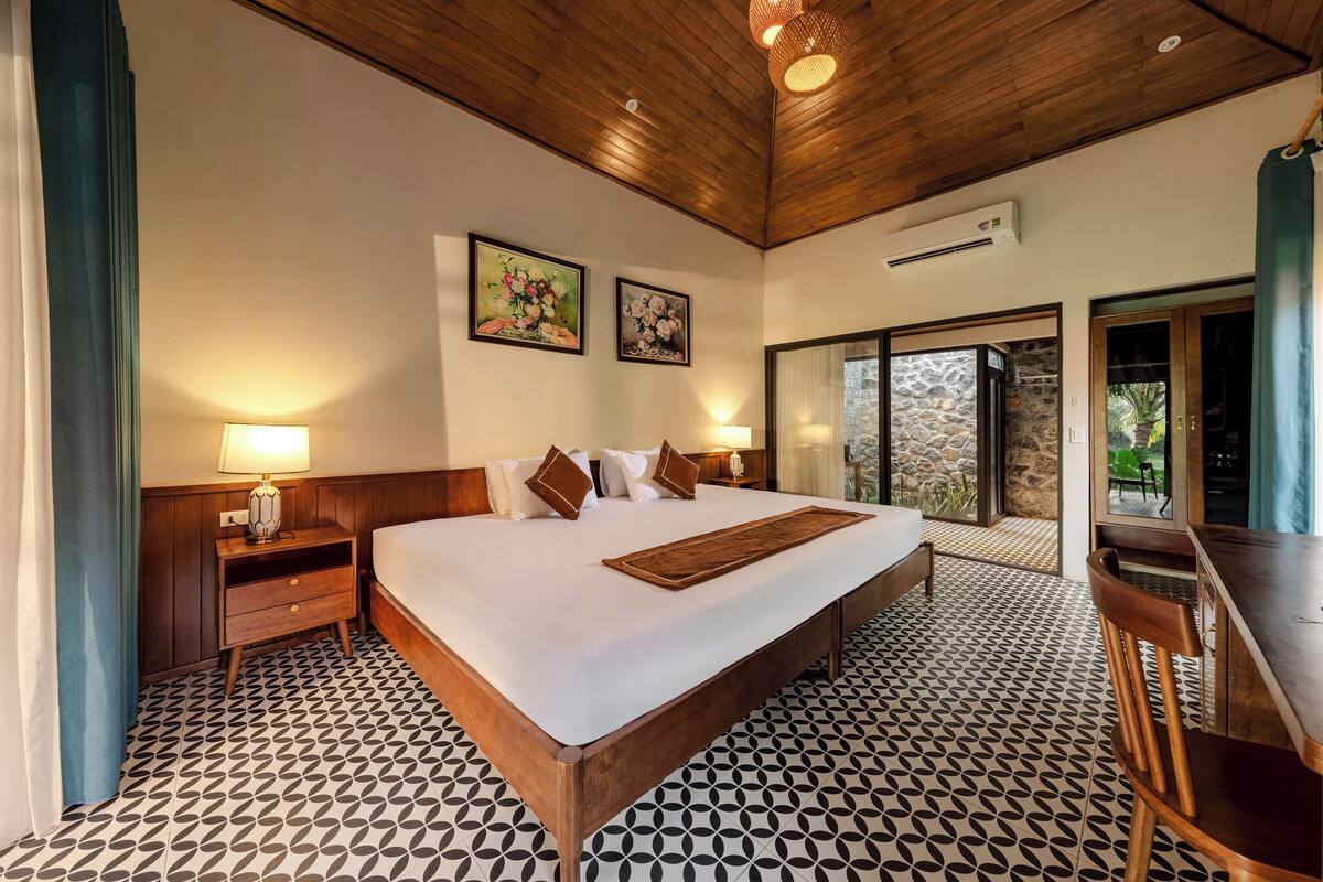 Habitación de hotel con una cama grande, cabecera de madera, dos cuadros en la pared, una mesita de noche con lámpara, un escritorio con silla y vistas a un espacio exterior a través de una puerta de cristal.