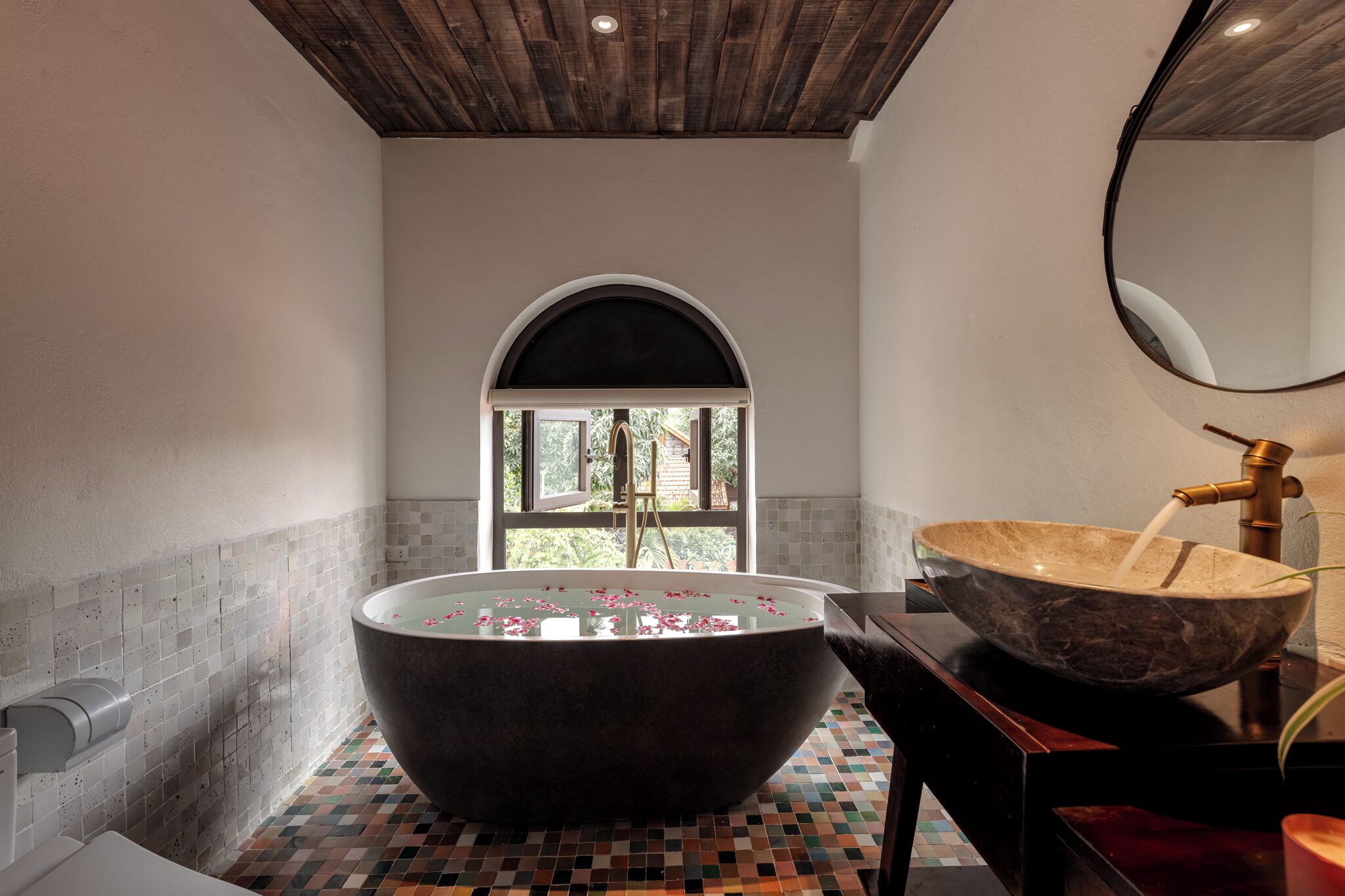 A modern bathroom with a freestanding bathtub, a large mirror, and a window with a view of greenery.