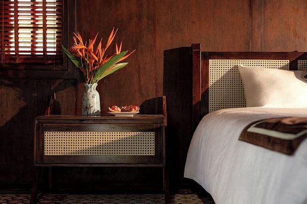 A wooden bed with a woven headboard, a bedside table with a vase of flowers, and a window with wooden blinds. - Nature Garden Ecolodge Ninh Binh (Tay Hoa Lu)
