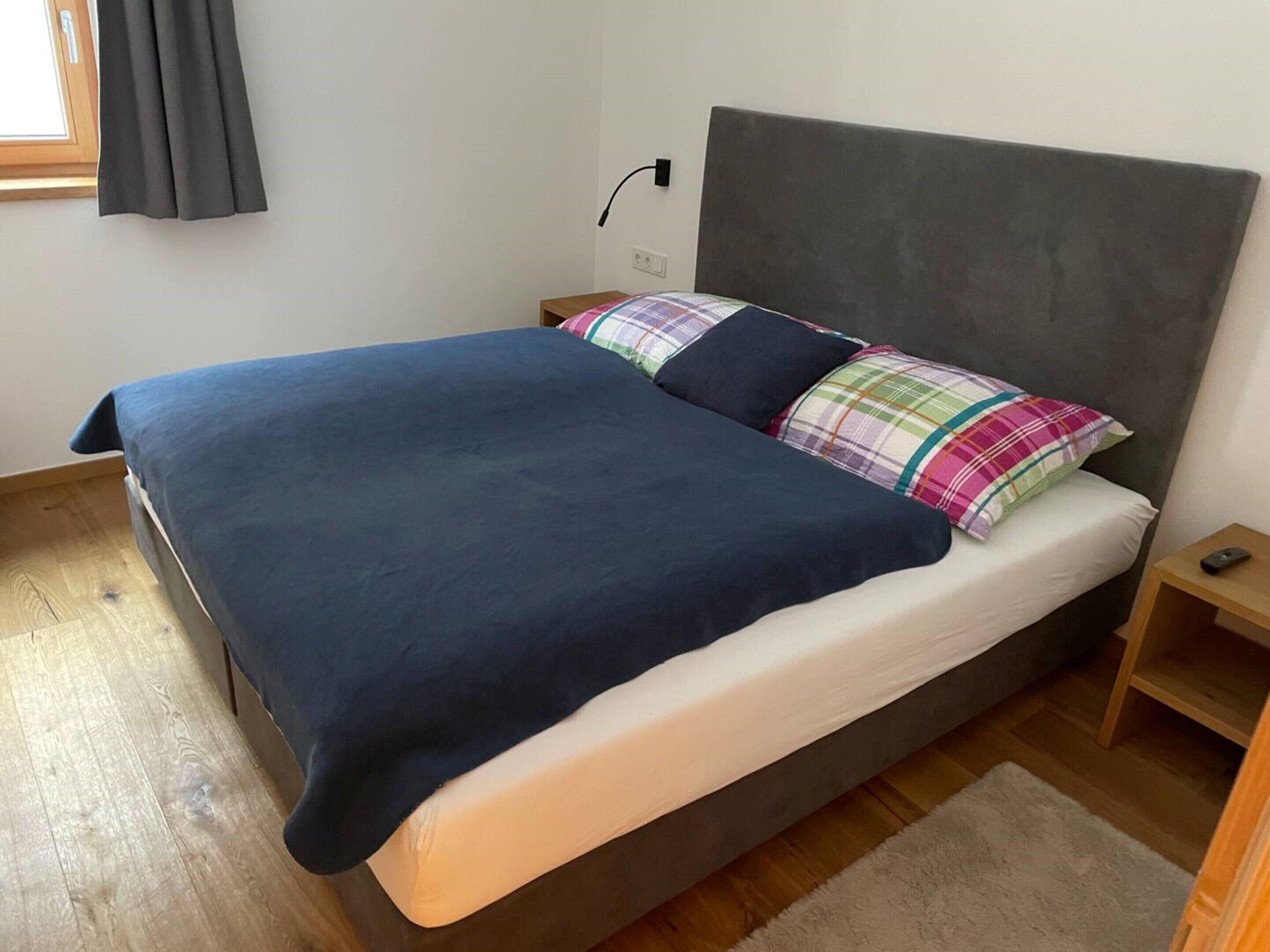 A bed with a dark blue blanket, a white mattress, and colorful pillows. There is a wooden nightstand with a remote control beside the bed.