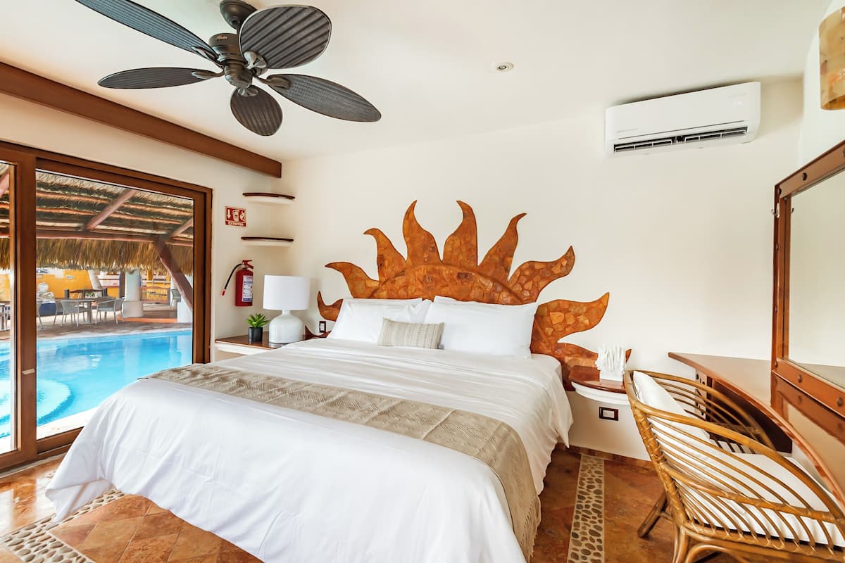A bedroom with a large bed, a ceiling fan, an air conditioning unit, a wooden headboard, a nightstand, a lamp, and a view of an outdoor pool.